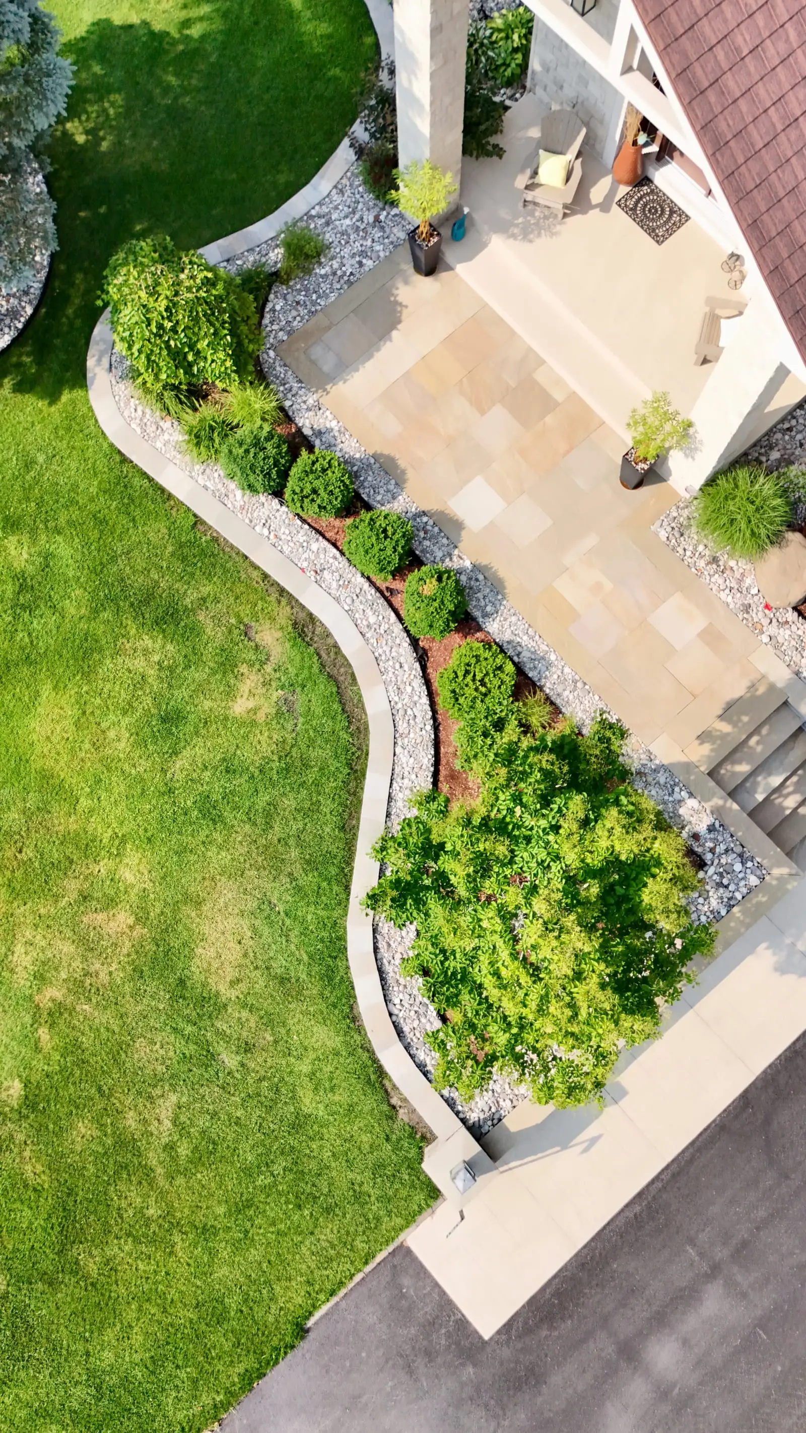 The curved bed from above — layered shrubs set in river-rock mulch behind a cut-stone edge, following the line of the sandstone patio