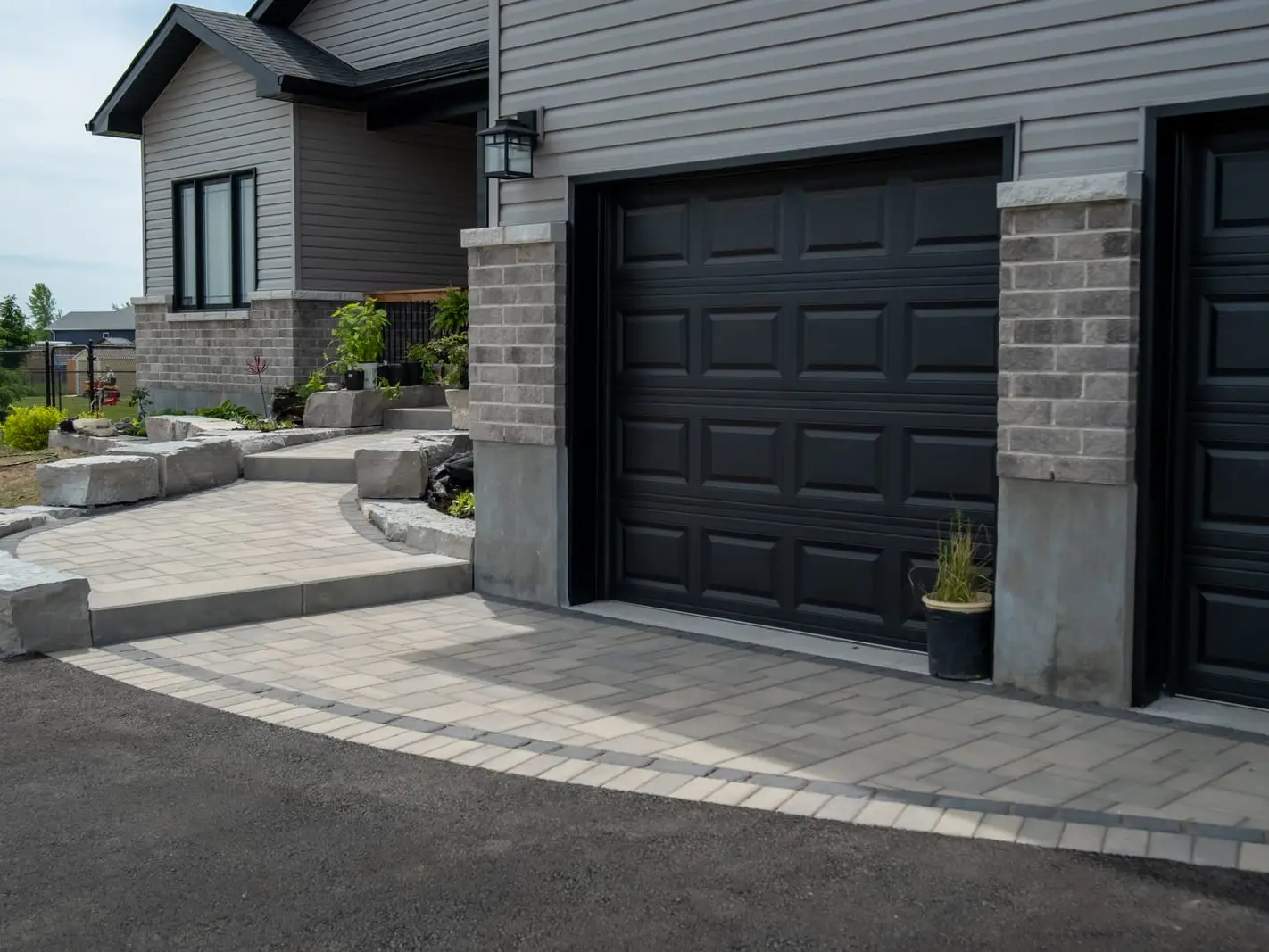 A pale interlock driveway edged with a contrasting soldier course, curving past armour-stone steps that climb to the front walk