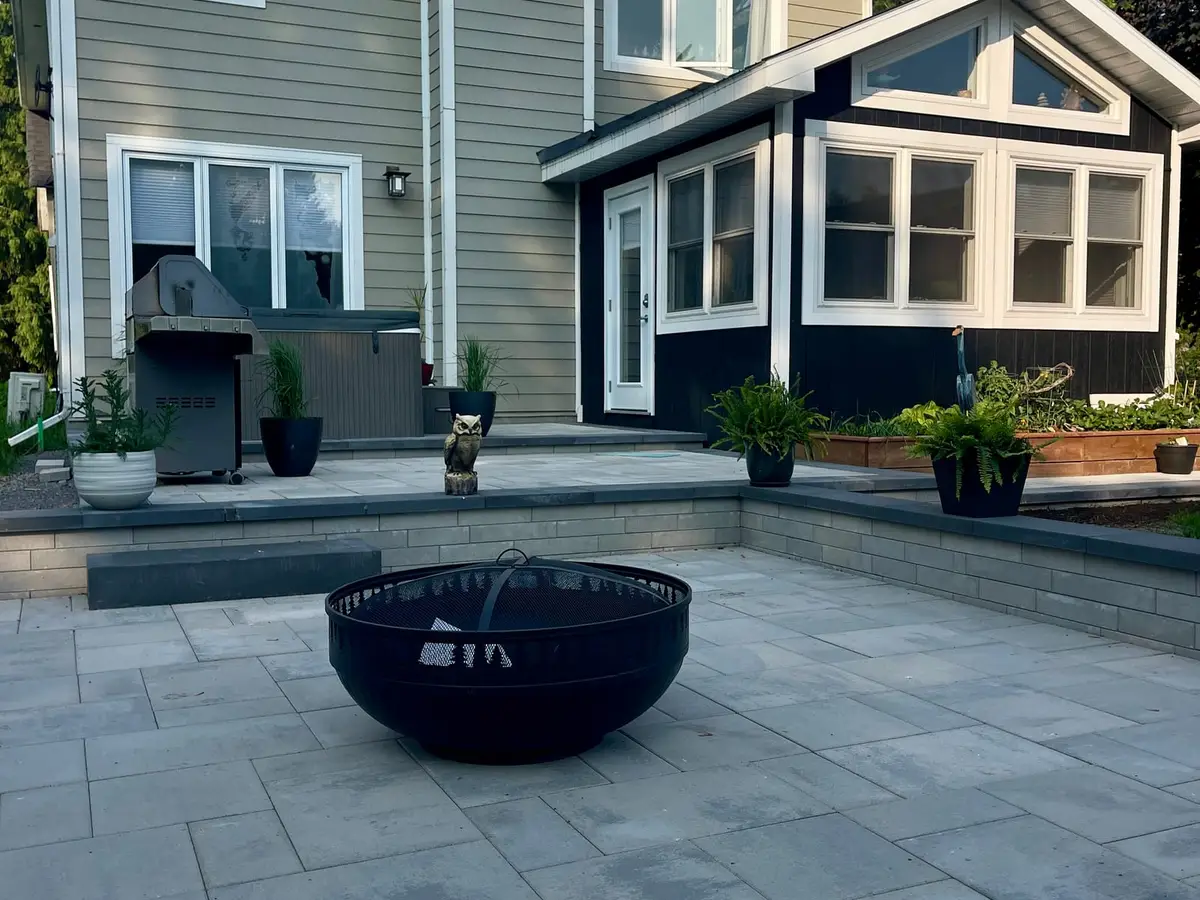 A grey slab terrace in evening light with a black steel fire bowl at its centre, charcoal-edged steps rising to an upper tier where a barbecue and hot tub sit against the house