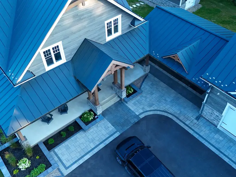 Aerial view of a home's front entrance with an interlock paver walkway and charcoal border band wrapping the driveway, and stone-edged garden beds along the porch